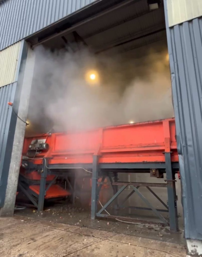 White water mist from a high pressure odor suppression line at the mouth of an industrial hall, above an orange conveyor